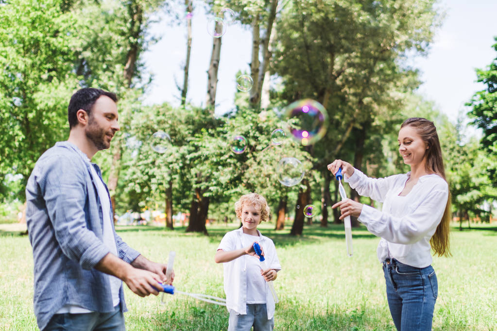 Mutter, Vater und Sohn in Freizeitkleidung spielen im Park mit Seifenblasen - Foto, Bild