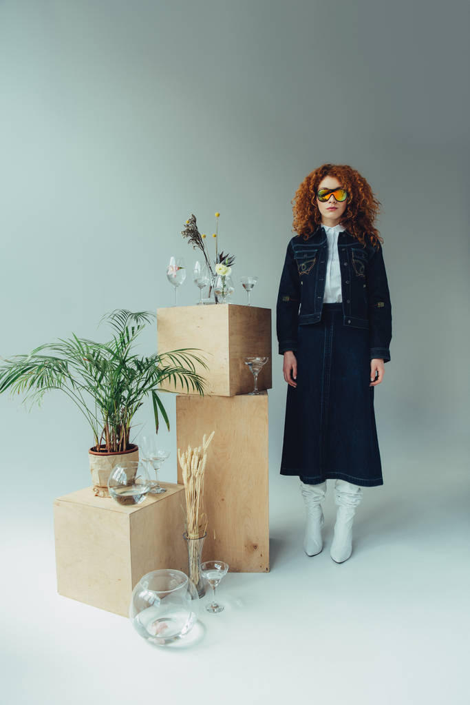 ragazza rossa alla moda in posa vicino a scatole di legno, occhiali e piante su grigio
 - Foto, immagini