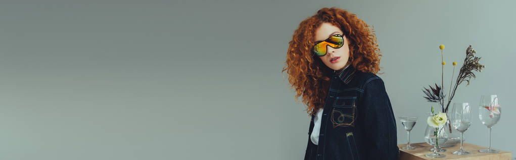 panoramic shot of stylish redhead girl in sunglasses posing near glasses and plants  isolated on grey - Photo, Image