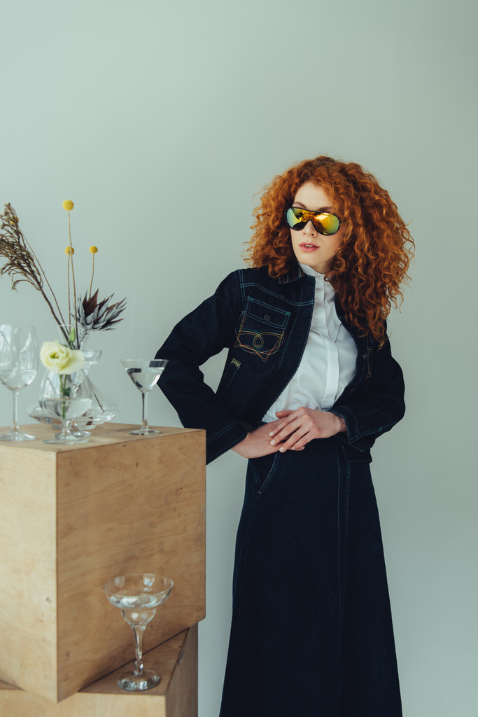 chica pelirroja de moda posando cerca de cajas de madera, gafas y plantas aisladas en gris
 - Foto, Imagen