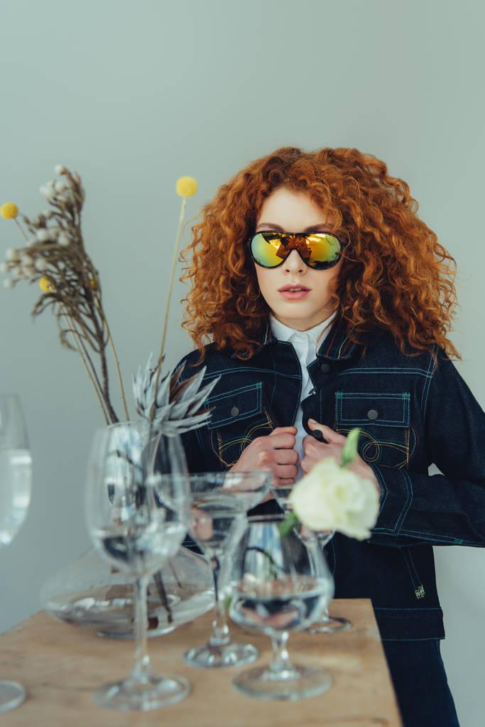 κομψή κοκκινομάλλα γυναίκα σε γυαλιά ηλίου που θέτουν κοντά γυαλιά και αποξηραμένα λουλούδια απομονώνονται σε γκρι - Φωτογραφία, εικόνα