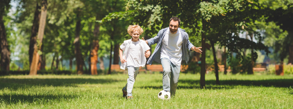 panoramatický záběr otce a roztomilého syna, který hrál fotbal s fotbalovým míčkem v parku - Fotografie, Obrázek
