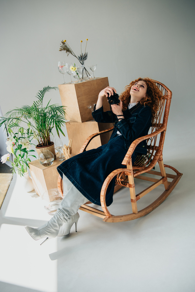 stylish laughing girl in wicker chair with film camera near wooden boxes, glasses and plants on grey - Photo, Image