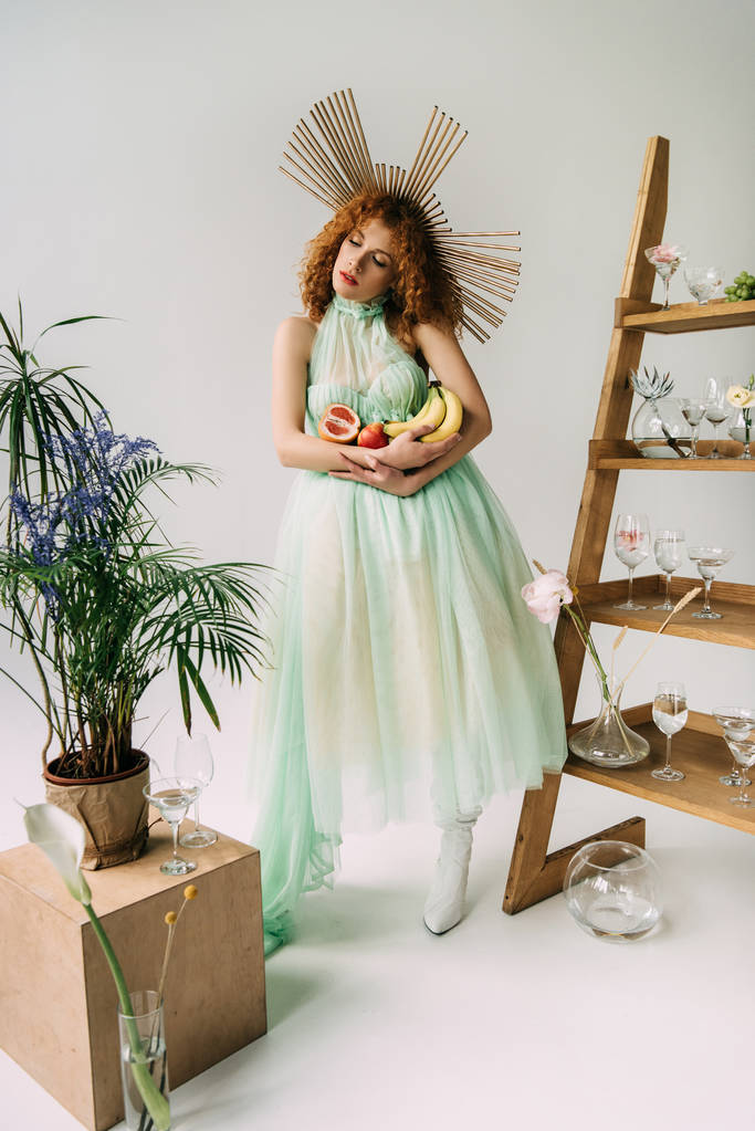 stylish redhead girl with accessory on head and fruits near ladder with flowers and glasses - Photo, Image