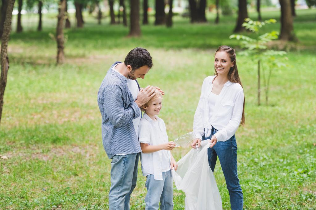 family in casual clothes picking up garbage in plastic bag in park - Photo, Image
