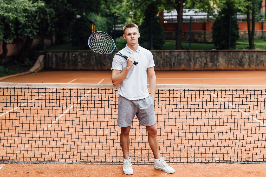 Knappe jongeman in polo shirt met tennisracket en glimlach terwijl hij op de tennisbaan staat - Foto, afbeelding