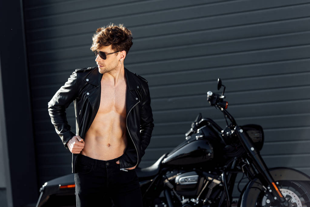 mladý muž se svalovým trupem v kožené bundě stojící blízko černého motocyklu - Fotografie, Obrázek