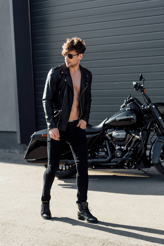 молодой человек в солнечных очках и кожаной куртке, идущий рядом с черным мотоциклом
 - Фото, изображение