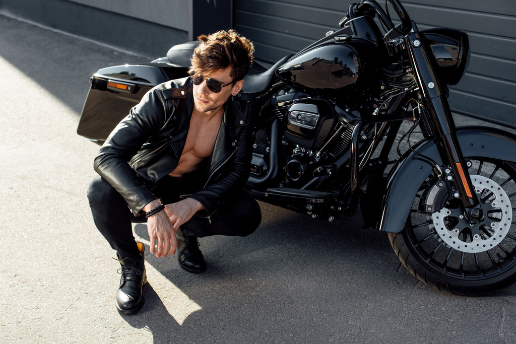 joven musculoso sentado cerca de la motocicleta y mirando hacia otro lado
 - Foto, Imagen