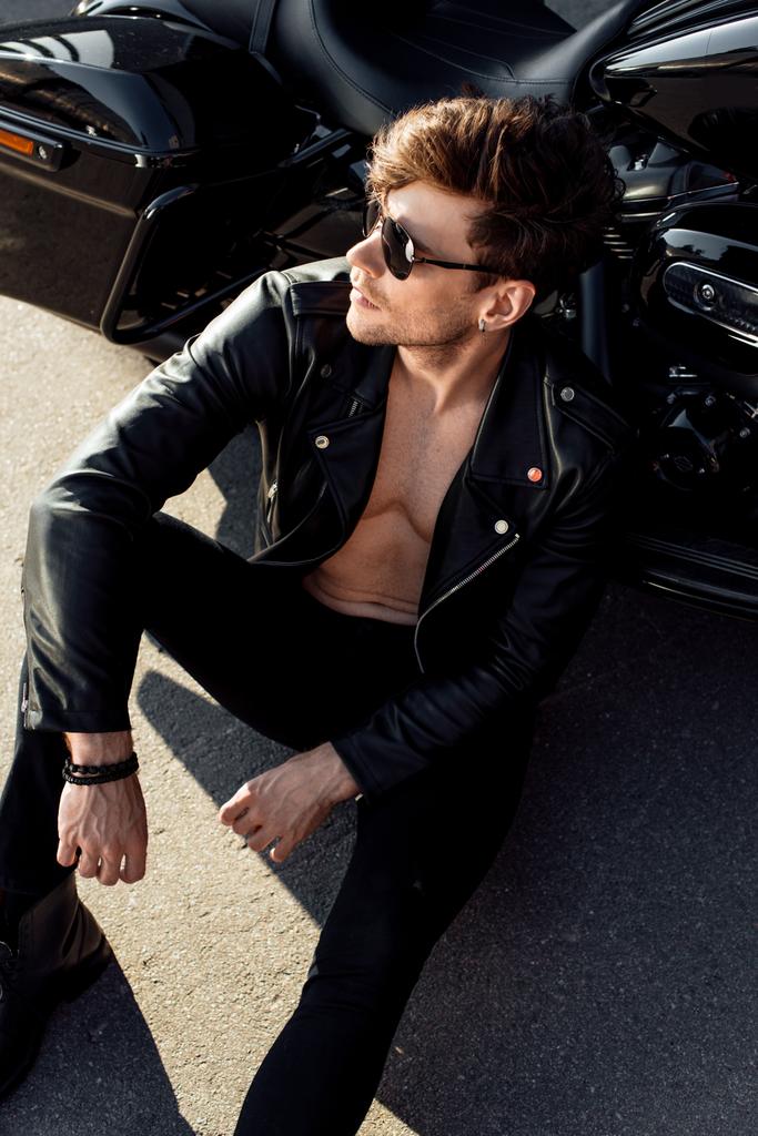 schöner Mann mit muskulösem Oberkörper sitzt auf dem Boden neben Motorrad und schaut weg - Foto, Bild