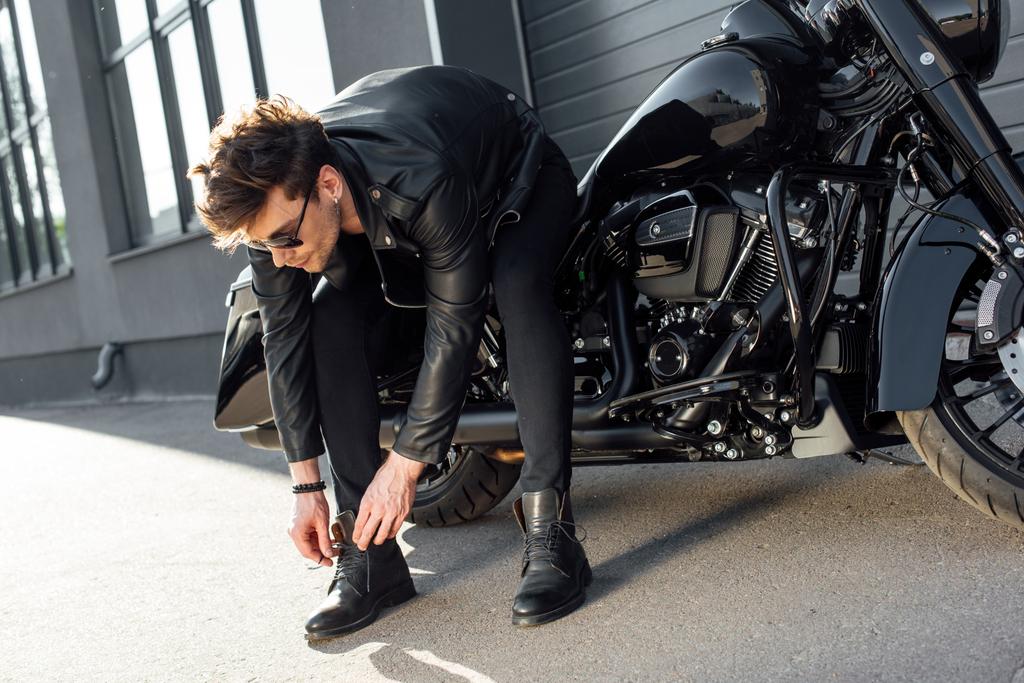 jonge man in lederen jas vetersluiting laarzen terwijl zittend op zwarte motorfiets - Foto, afbeelding