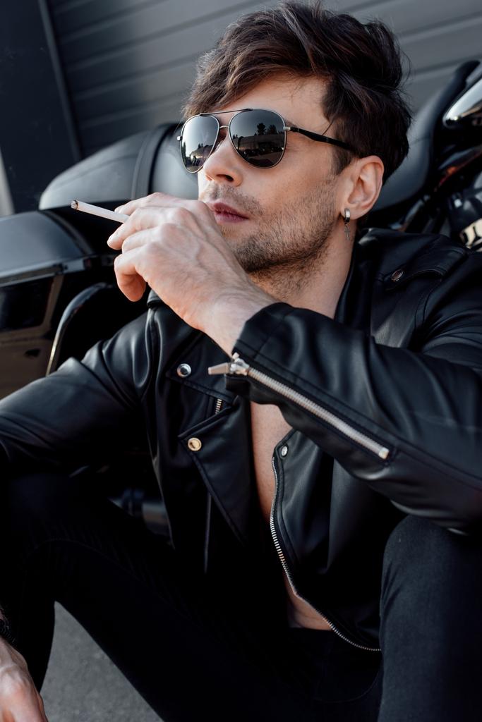 joven con gafas de sol y chaqueta de cuero sosteniendo el cigarrillo mientras está sentado cerca de la motocicleta
 - Foto, Imagen