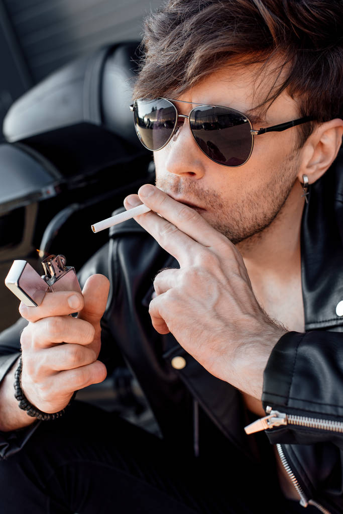 Portraitaufnahme eines jungen Mannes mit Sonnenbrille, der sich Zigarette anzündet, während er in der Nähe eines schwarzen neuen Motorrads sitzt - Foto, Bild