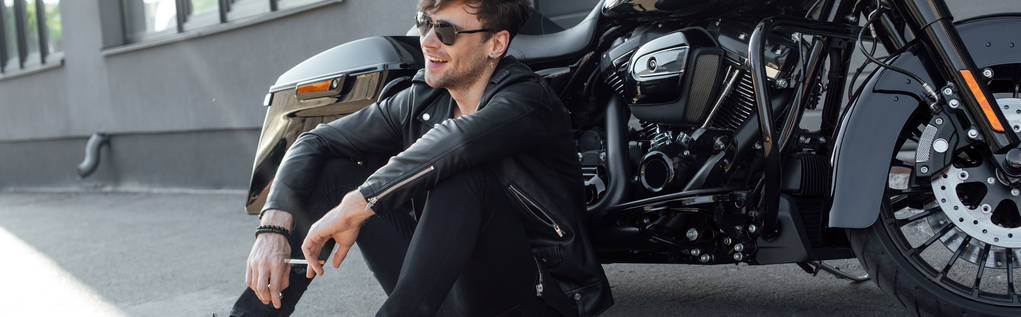 tiro panorâmico de jovem sentado perto de motocicleta preta, sorrindo e segurando cigarro
 - Foto, Imagem
