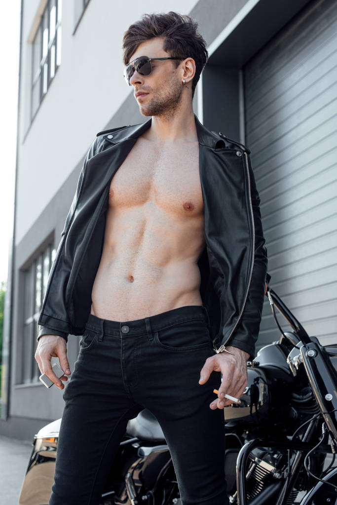 junger Motorradfahrer mit muskulösem nacktem Oberkörper steht neben schwarzem Motorrad, schaut weg und hält Zigarette - Foto, Bild