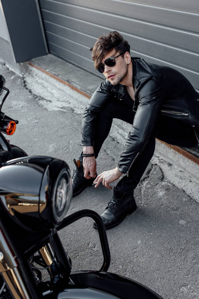 handsome motorcyclist in sunglasses sitting on ground near motorcycle - Photo, Image