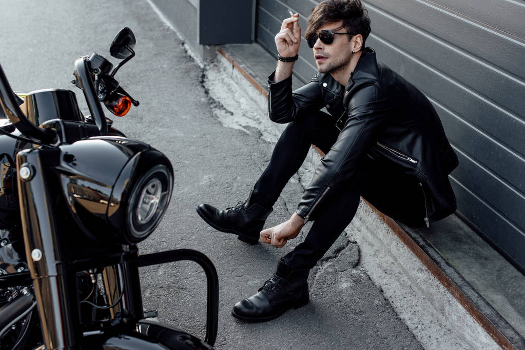 bello motociclista in occhiali da sole seduto a terra vicino alla moto e fumare sigaretta
  - Foto, immagini