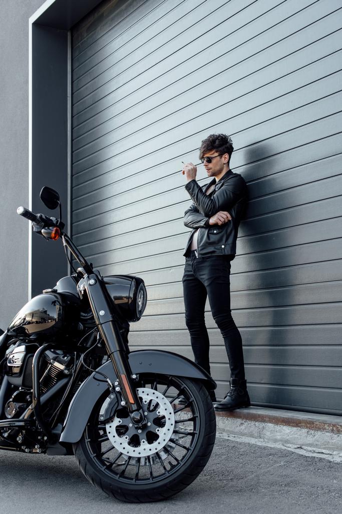 オートバイの近くに壁の近くに立って、タバコを保持するサングラスでハンサムなモーターサイクリストの完全な長さのビュー  - 写真・画像