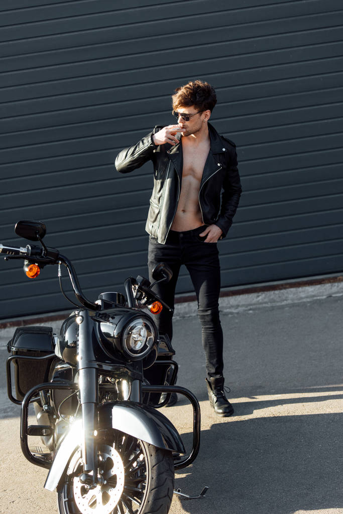 Ganzkörperansicht eines Motorradfahrers mit muskulösem Oberkörper, der neben einem Motorrad steht und Zigarette in der Hand hält  - Foto, Bild