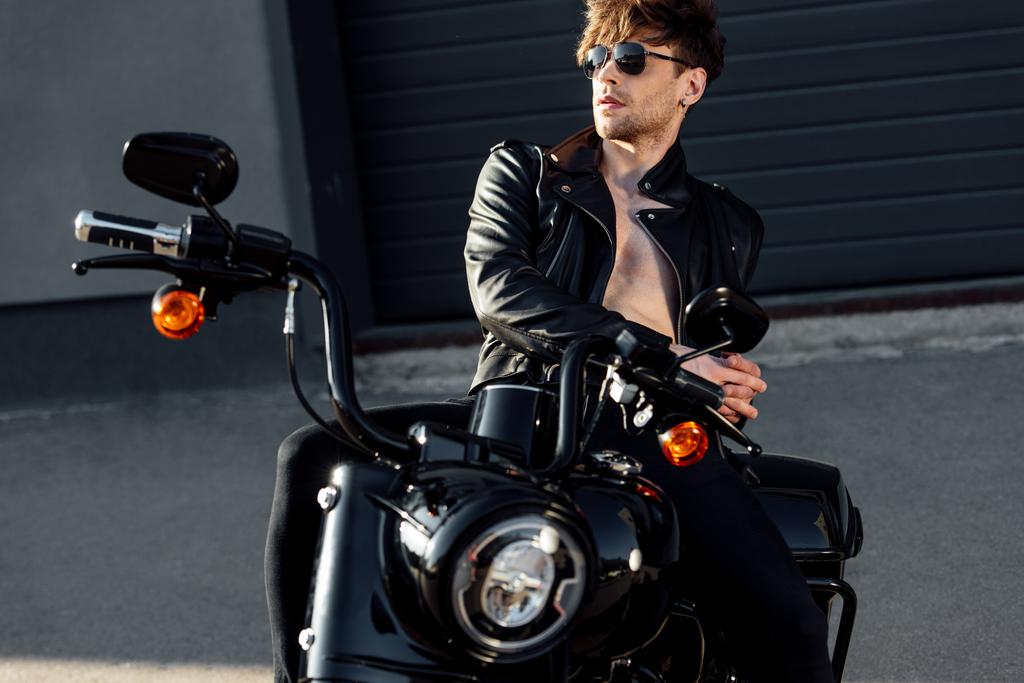 красивий молодий мотоцикліст з голим в сонцезахисних окулярах і шкіряний піджак торсо, що спирається на чорний мотоцикл
  - Фото, зображення