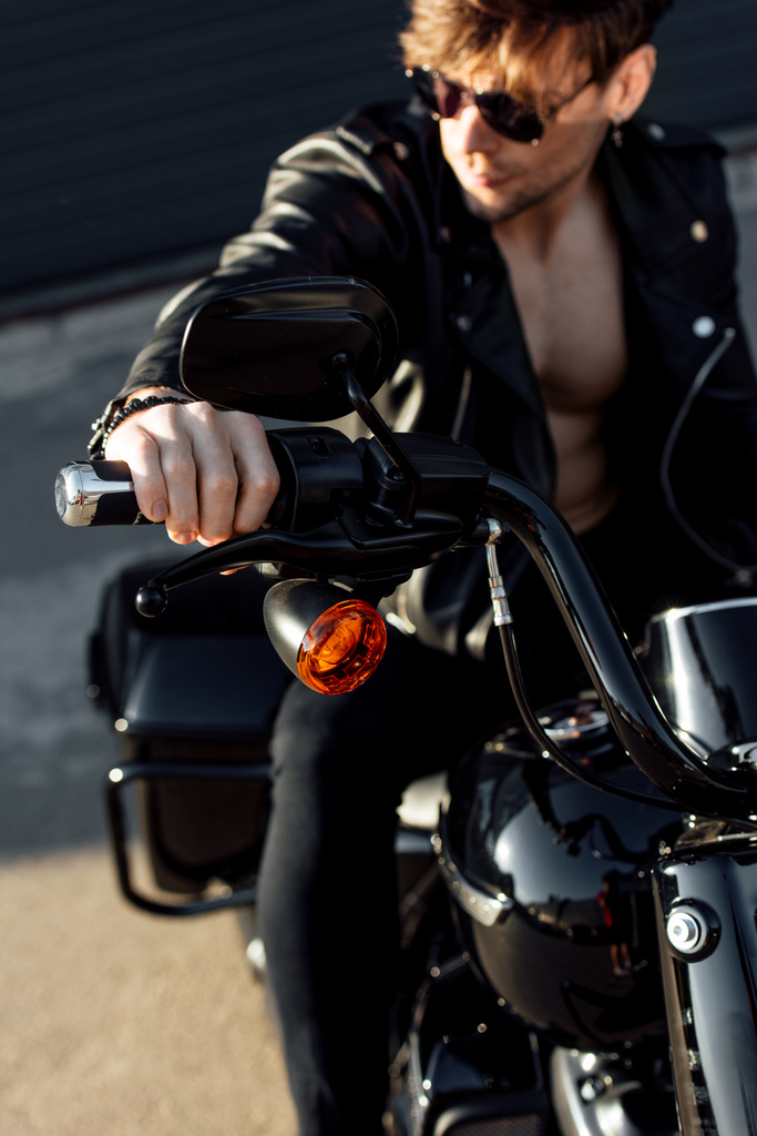 selectieve focus van jonge man zittend op motorfiets in zonlicht, vasthouden handvat en wegkijken - Foto, afbeelding