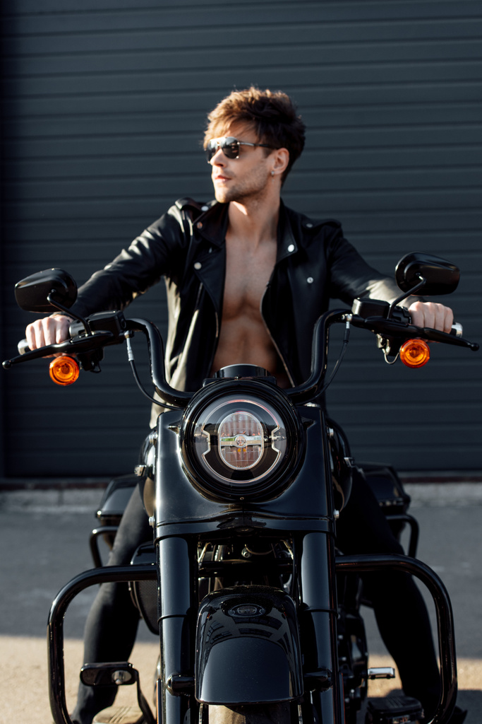guapo motociclista con torso desnudo muscular en chaqueta de cuero sentado en motocicleta y agarraderas
  - Foto, imagen
