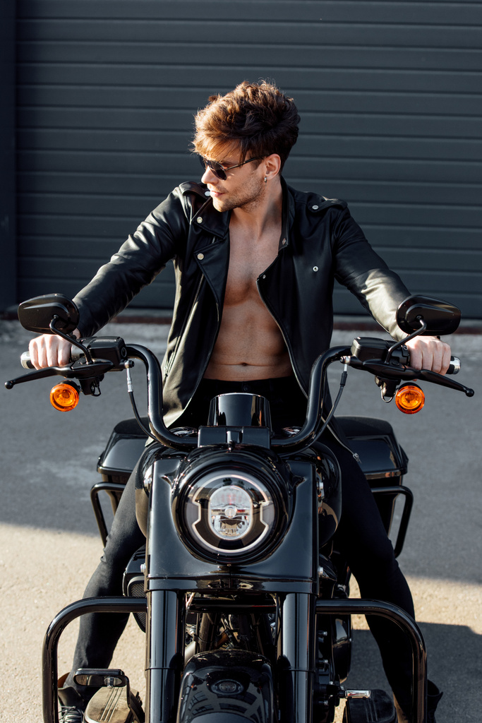 przystojny motocyklista z muskularnym nagi tułowia w skórzanej kurtce siedzącej na motocyklu, patrząc z dala i trzymając uchwyty  - Zdjęcie, obraz
