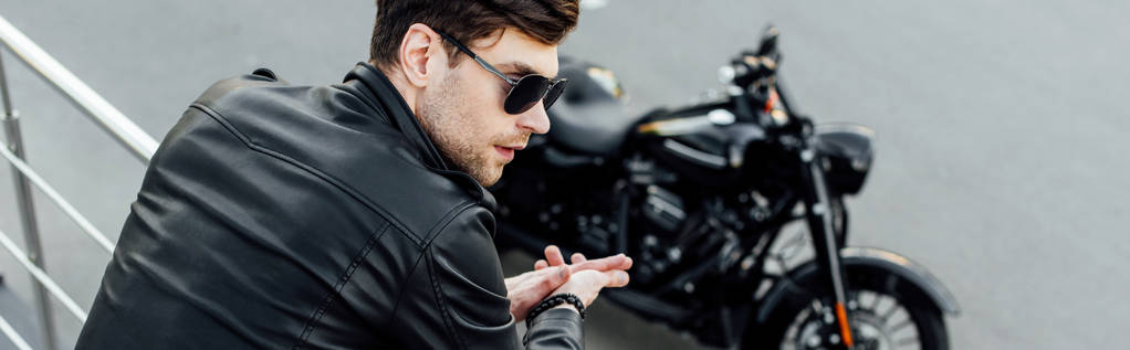 панорамний знімок молодого чоловіка в чорній шкіряній куртці, що стоїть біля металевого паркану з мотоциклом на фоні
 - Фото, зображення