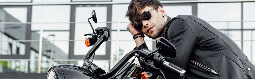 tiro panorámico de joven guapo en chaqueta de cuero descansando mientras está sentado en la motocicleta
  - Foto, imagen
