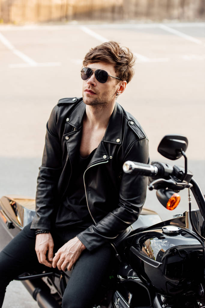 motocyklista w skórzanej kurtce siedzącej na czarnym motocyklu i patrząc od - Zdjęcie, obraz