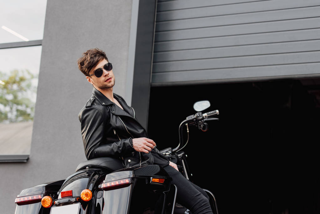 motocyklista w okulary przeciwsłoneczne i Kurtka skórzana siedzi na motocyklu w pobliżu otwartego garażu i patrząc na kamerę  - Zdjęcie, obraz