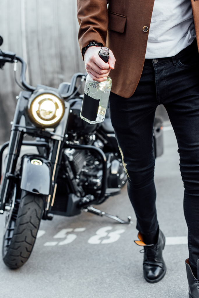 Ausgeschnittener Blick auf Mann in brauner Jacke, der neben Motorrad läuft und Alkoholflasche in der Hand hält  - Foto, Bild