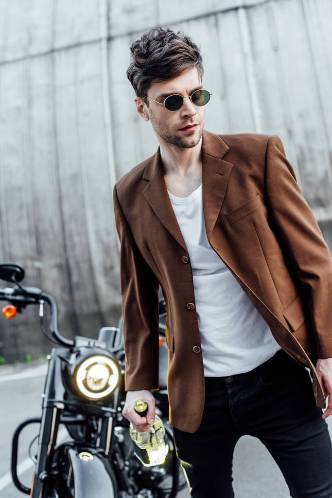 motosiklet yakınında alkol şişesi ile yürüyüş güneş gözlüğü ve kahverengi ceket yakışıklı adam seçici odak  - Fotoğraf, Görsel