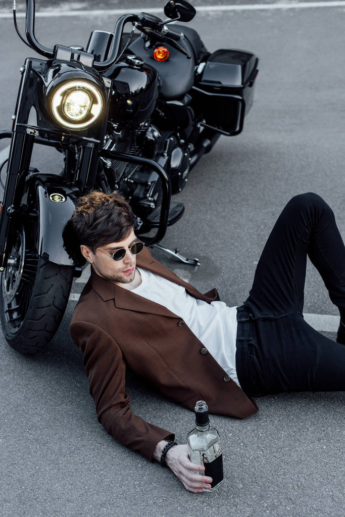 przystojny młody człowiek w okulary leżące na ziemi w pobliżu motocykla z świetlnej lampy  - Zdjęcie, obraz