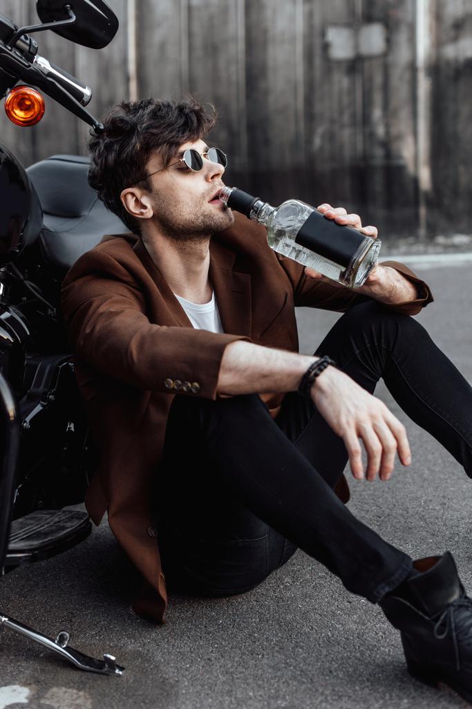 молодий чоловік п'є алкоголь, сидячи на землі біля мотоцикла
 - Фото, зображення