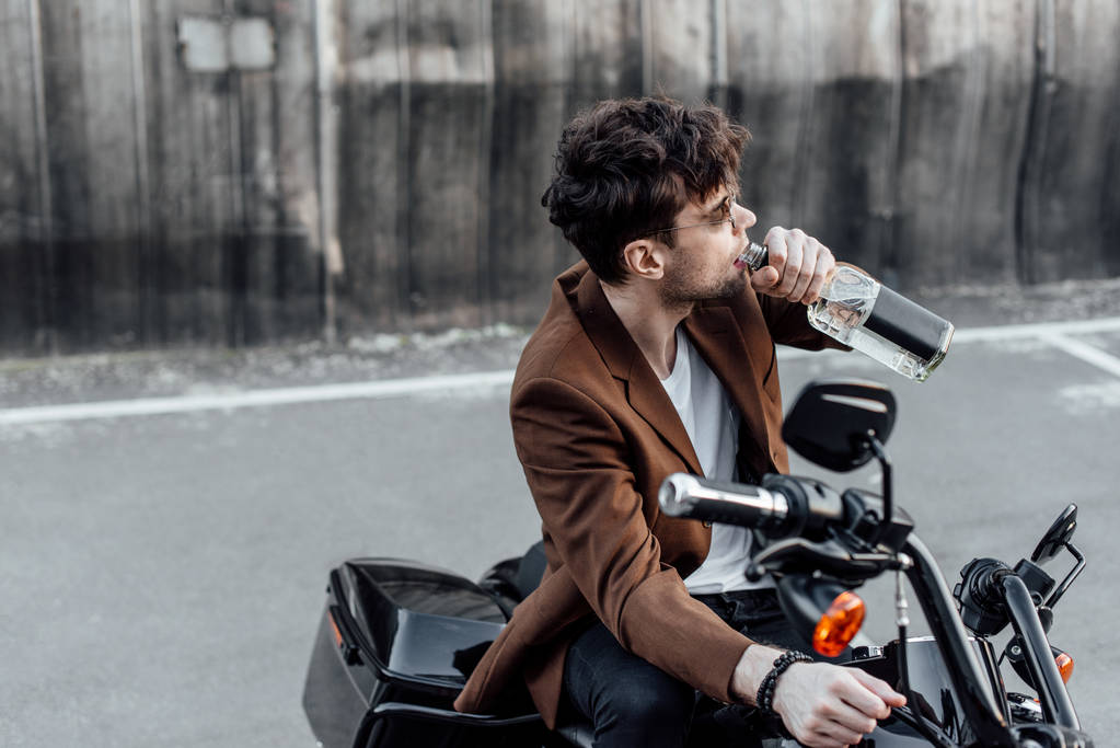 άνθρωπος σε καφέ μπουφάν πίνοντας αλκοόλ από το μπουκάλι ενώ κάθονται σε μοτοσικλέτα - Φωτογραφία, εικόνα