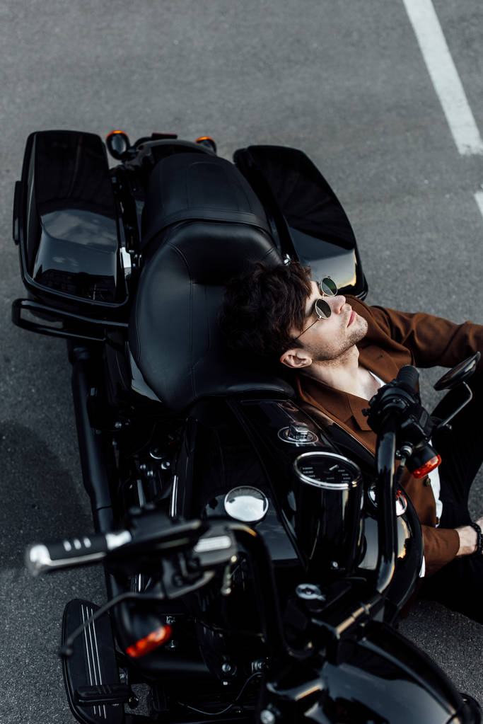 молодий чоловік в окулярах і коричневій куртці відпочиває, сидячи на землі і нахилившись на мотоциклі
 - Фото, зображення