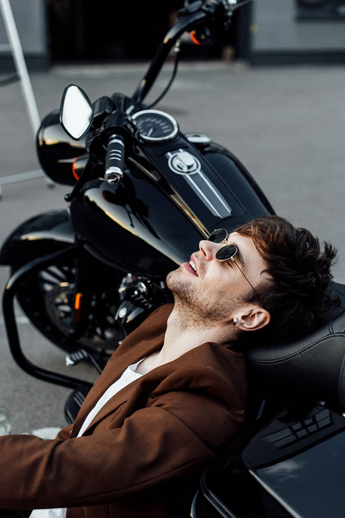 όμορφος μοτοσικλετιστής σε καφέ μπουφάν αναπαύσεως ενώ γέρνει το κεφάλι σε μοτοσικλέτα - Φωτογραφία, εικόνα