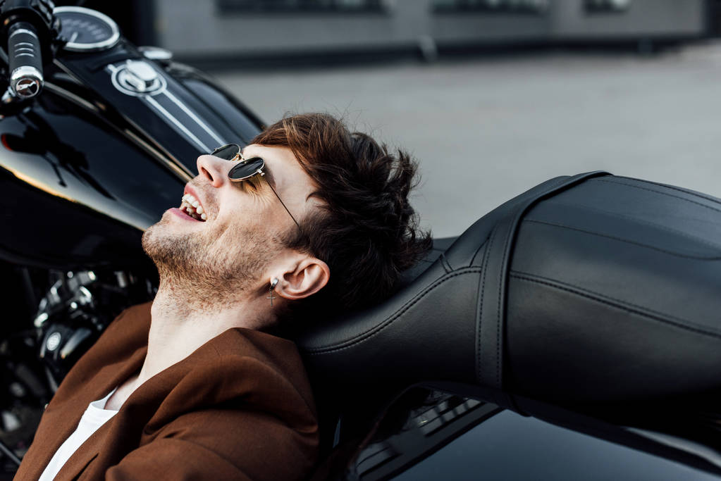 szczęśliwy młody człowiek w brązowej kurtki uśmiechnięty i odpoczynku podczas pochylony głowę na motocyklu  - Zdjęcie, obraz