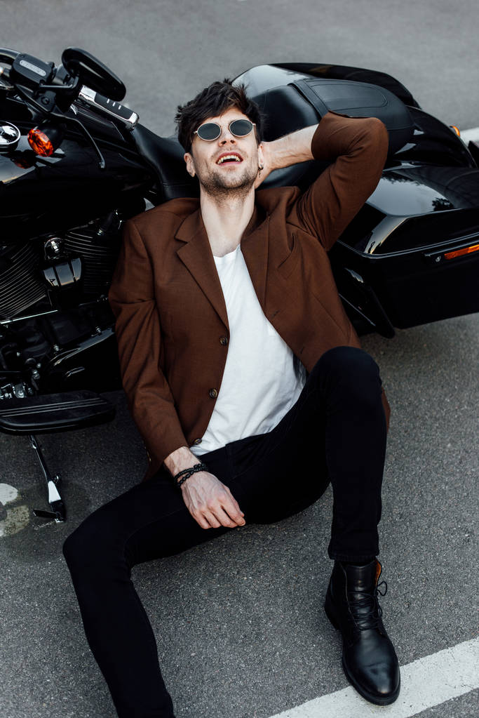 handsome man sitting on ground smiling and resting while leaning head on motorcycle  - Photo, Image