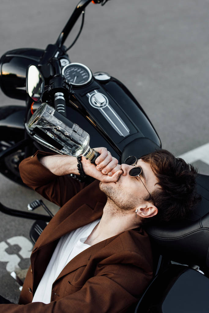 κομψό μοτοσικλετιστής που γέρνει το κεφάλι σε μοτοσικλέτα και πόσιμο αλκοόλ  - Φωτογραφία, εικόνα