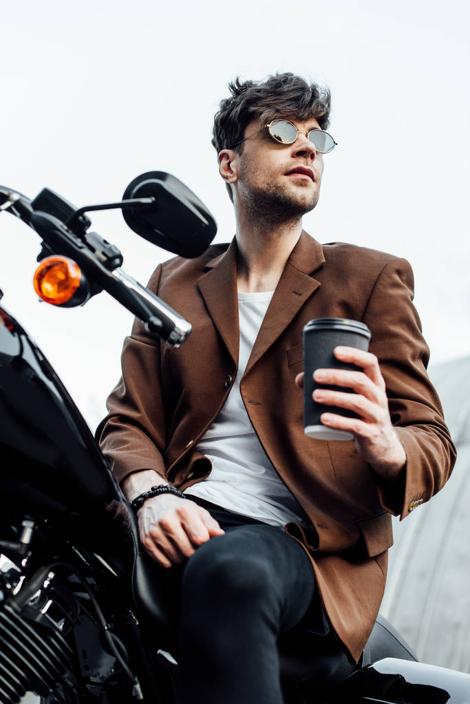 オートバイに座っている間に行くためにコーヒーを保持するジャケットとサングラスでスタイリッシュな男の低角度のビュー - 写真・画像