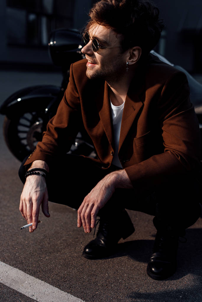 молодой человек в солнцезащитных очках и куртке сидит на корточках возле мотоцикла и курит сигарету
  - Фото, изображение