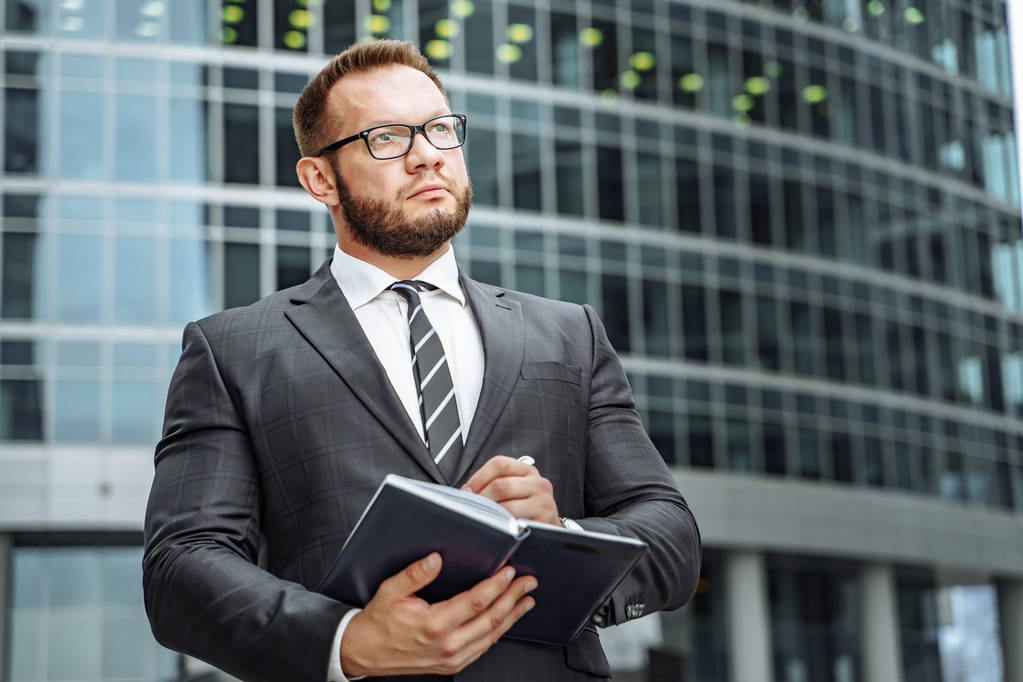 Porträt eines erfolgreichen Geschäftsmannes in Anzug und Brille mit einem Notizbuch in der Hand auf dem Hintergrund des Business Centers - Foto, Bild