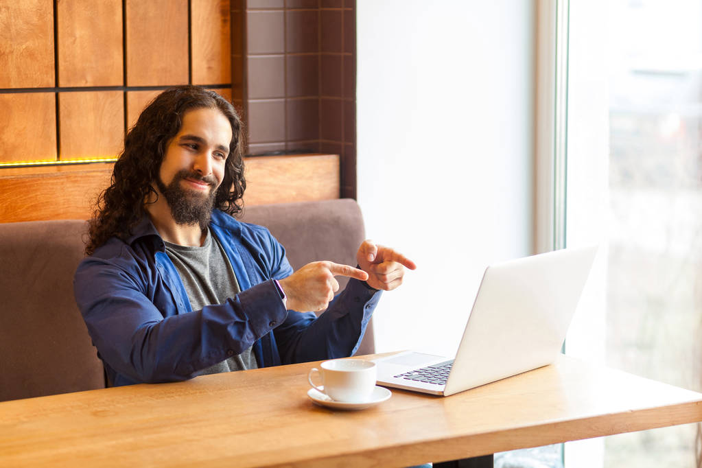 joyeux beau jeune homme adulte pigiste dans un style décontracté assis dans un café et parler avec un ami dans un ordinateur portable tout en montrant deux doigts à l'écran
  - Photo, image