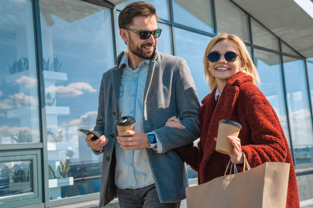 Beautiful young couple in sunglasses looking away and smiling while going on city street after shopping with coffee and mobile phone in hands - Photo, Image