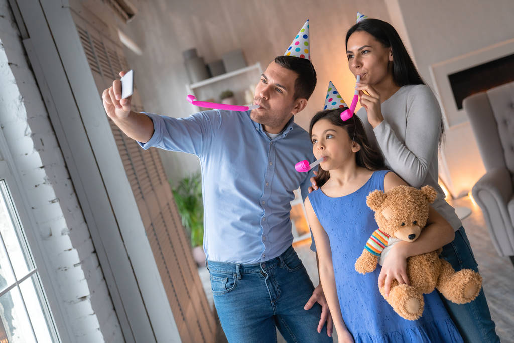 Ευτυχισμένη οικογένεια γιορτάζει τα γενέθλιά της μαζί στο σπίτι. Άνθρωπος που παίρνει selfie στο smartphone με ελκυστική γυναίκα μυρμήγκι χαριτωμένη μικρή κόρη τους - Φωτογραφία, εικόνα