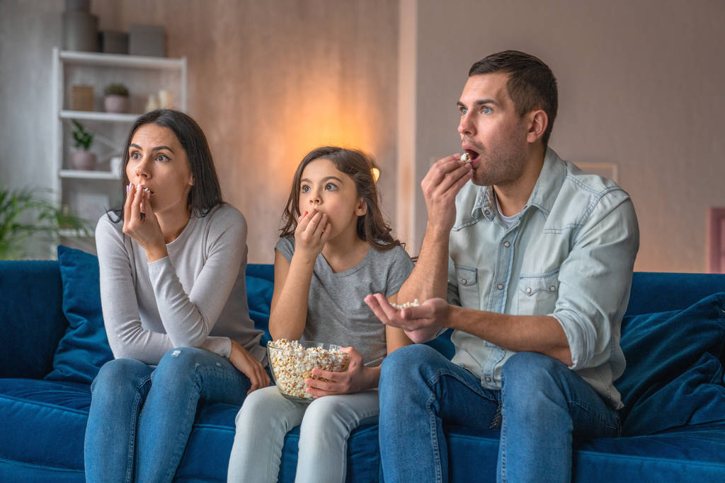 Возбужденные молодые родители и их милая маленькая дочь смотрят кино по телевизору, едят попкорн и сидят дома на диване
 - Фото, изображение