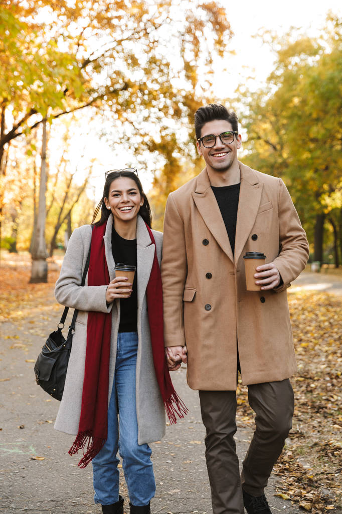 Porträt eines lässigen Paares, das Kaffee zum Mitnehmen trinkt und beim Spaziergang im Herbstpark in die Kamera lächelt - Foto, Bild