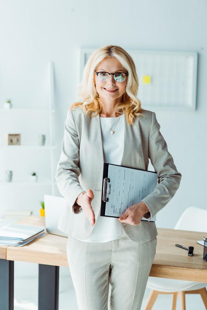 aantrekkelijke blonde vrouw in glazen houden Klembord en gebaren in Office  - Foto, afbeelding